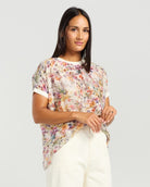 A woman modeling a short-sleeve, semi-sheer floral print top with white trim on the collar and sleeve cuffs, paired with white trousers, against a light gray studio background.