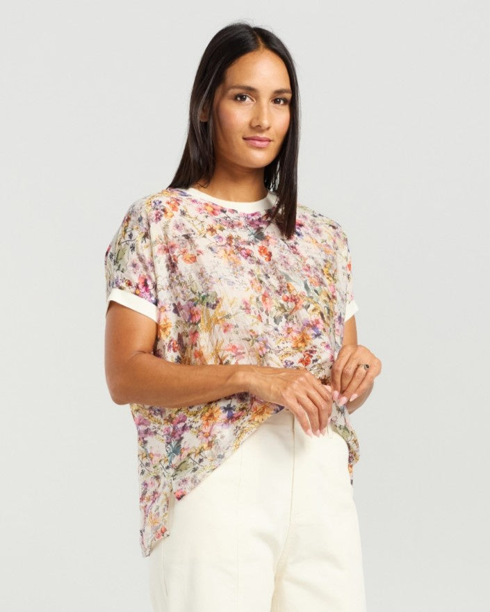A woman modeling a short-sleeve, semi-sheer floral print top with white trim on the collar and sleeve cuffs, paired with white trousers, against a light gray studio background.