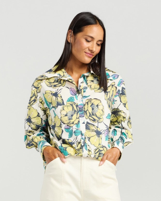 A woman wearing a long-sleeve button-up blouse with a vibrant yellow, green, and teal floral print on a white base, paired with cream-colored pants, set against a solid light gray background.