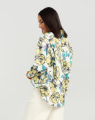 A woman shown from the back wearing a white button-up blouse with a large yellow and green floral print, paired with white trousers, against a light gray studio background.