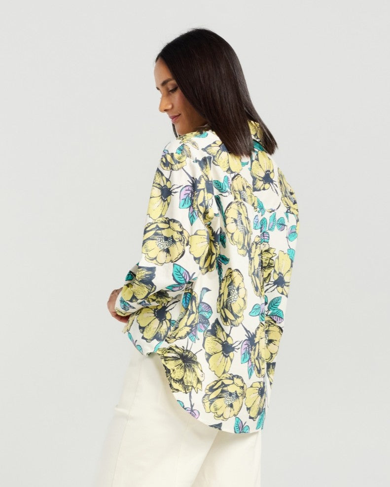 A woman shown from the back wearing a white button-up blouse with a large yellow and green floral print, paired with white trousers, against a light gray studio background.