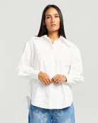 A woman wearing a white long-sleeve button-up shirt with a relaxed, oversized fit and blue jeans, standing against a light gray studio background.