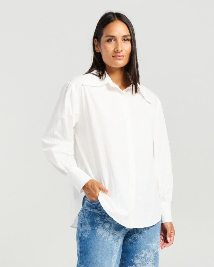 A woman modeling a white long-sleeve button-up blouse with a relaxed, oversized fit and a pointed collar, paired with blue jeans, against a light gray studio background.