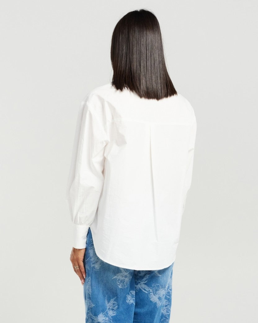 A woman shown from the back wearing a white long-sleeve blouse with gathered detailing and blue patterned pants, against a plain light gray studio background.