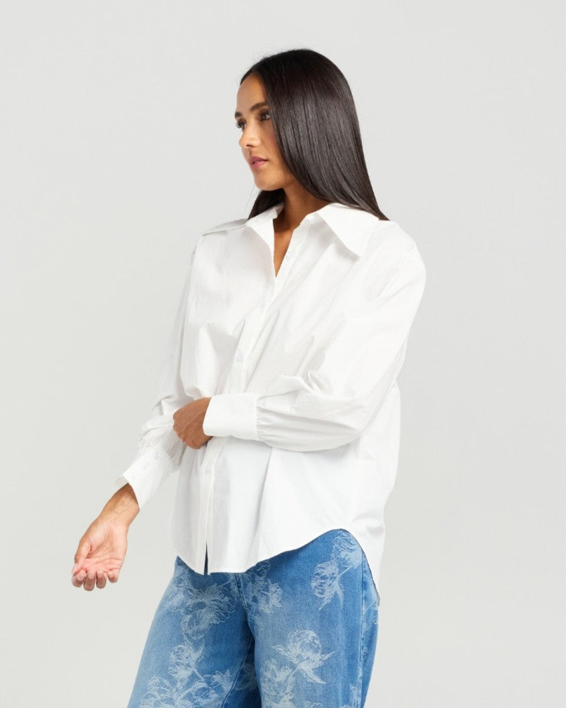 A woman wearing a white long-sleeve button-up shirt and blue floral-print jeans, photographed against a plain light gray studio background.