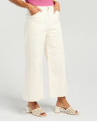 A model wearing off-white cropped wide-leg pants with a pink ribbed top and beige open-toe block-heel mules, shown from the waist down on a light gray studio background.