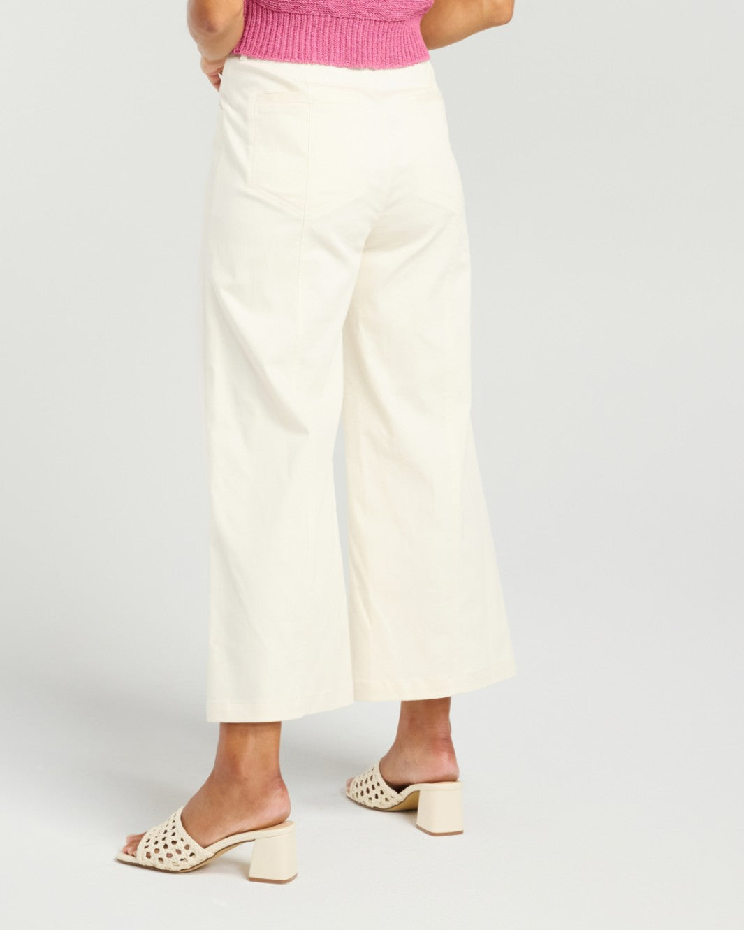 A person shown from the waist down wearing off-white cropped wide-leg pants, a pink ribbed top, and cream block-heel slide sandals against a light gray studio background.