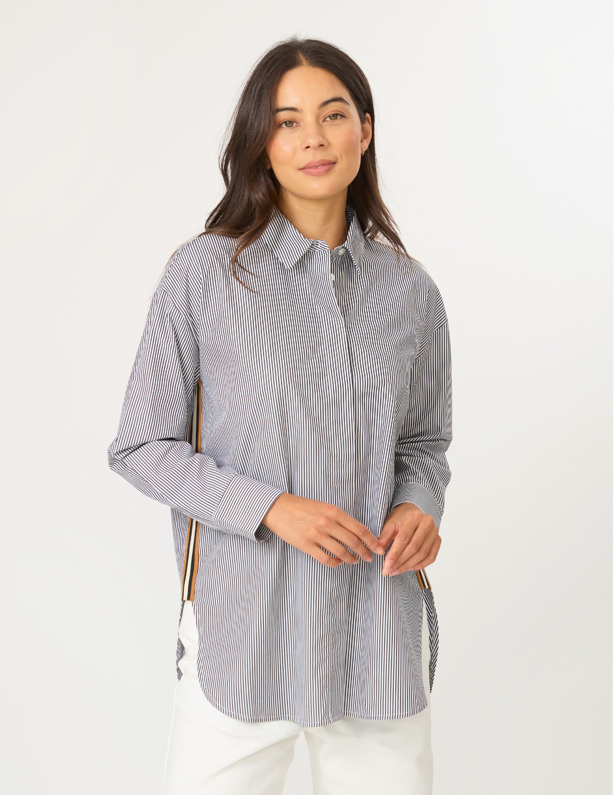 A woman modeling a light gray and white vertically striped button-down shirt with white pants, standing against a plain white studio background.