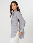 A woman wearing a light blue and white vertically striped button-down shirt with a distinctive multi-colored stripe detail on the sleeve, paired with white pants, against a plain light gray background.