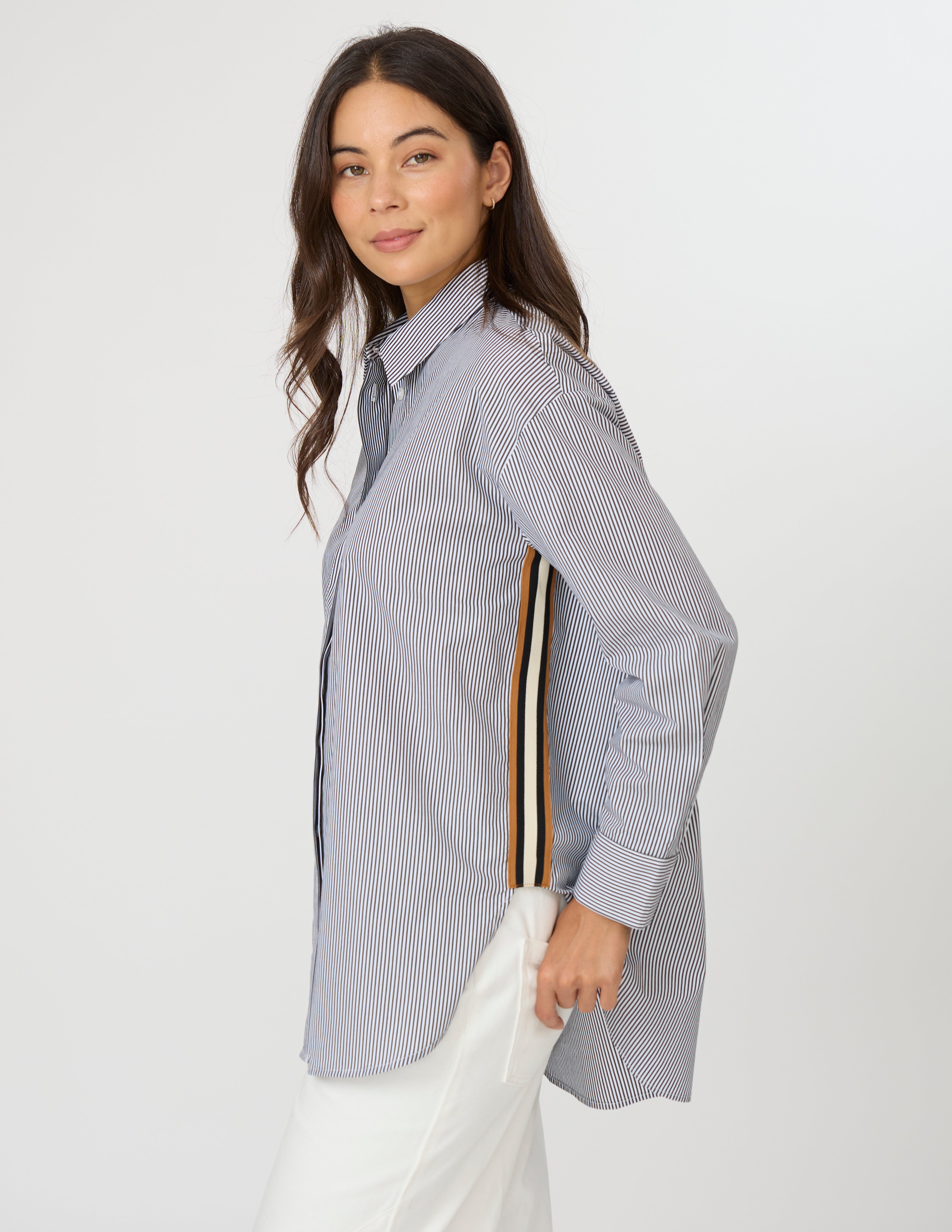 A woman wearing a light blue and white vertically striped button-down shirt with a distinctive multi-colored stripe detail on the sleeve, paired with white pants, against a plain light gray background.