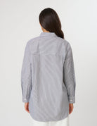 A woman viewed from the back wearing a light gray and white vertically striped button-up shirt with a large back pocket, set against a plain light gray studio background.