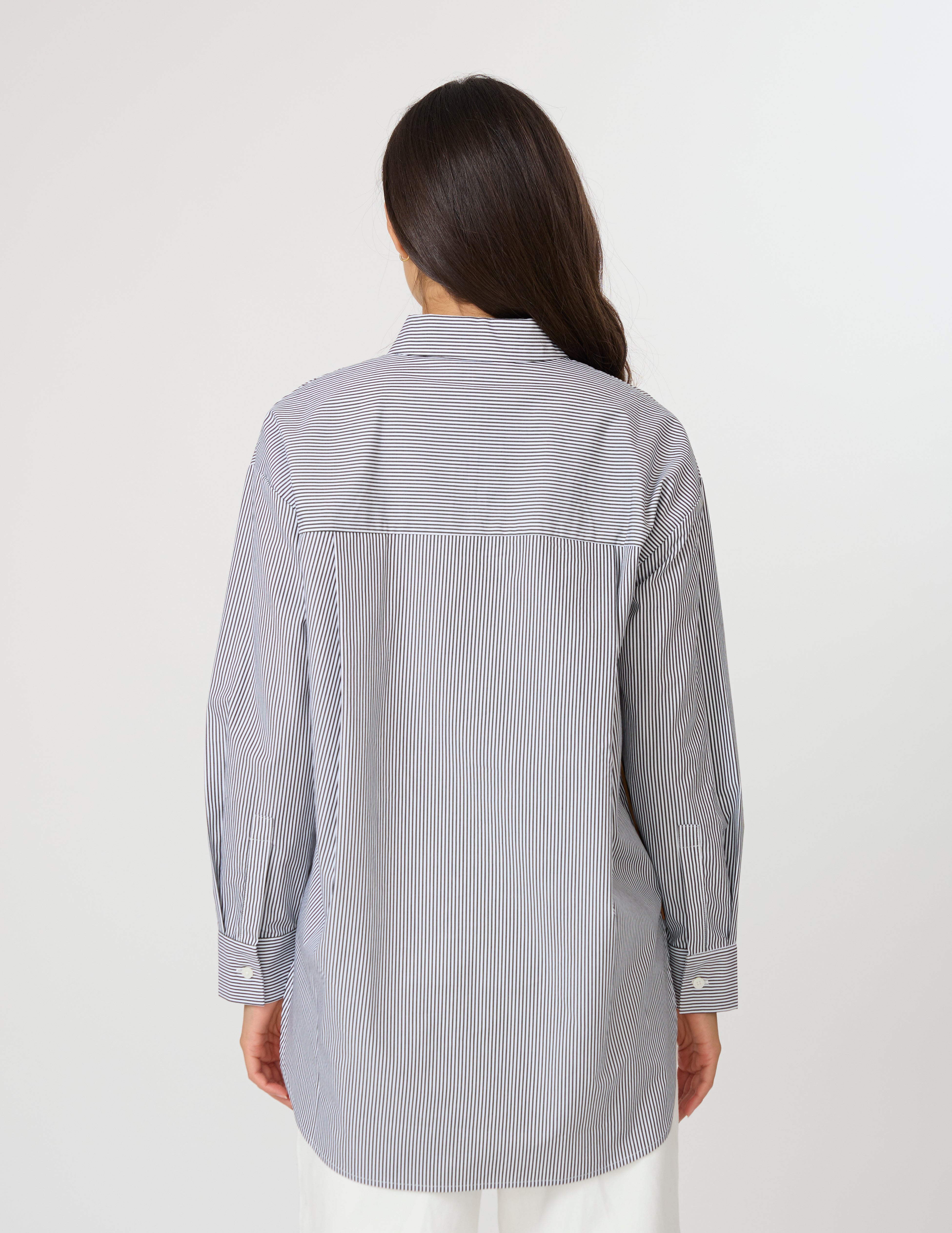A woman viewed from the back wearing a light gray and white vertically striped button-up shirt with a large back pocket, set against a plain light gray studio background.