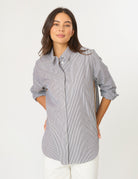 A woman modeling a light gray and white vertically striped button-down shirt with rolled-up sleeves, paired with white pants, against a plain white studio background.