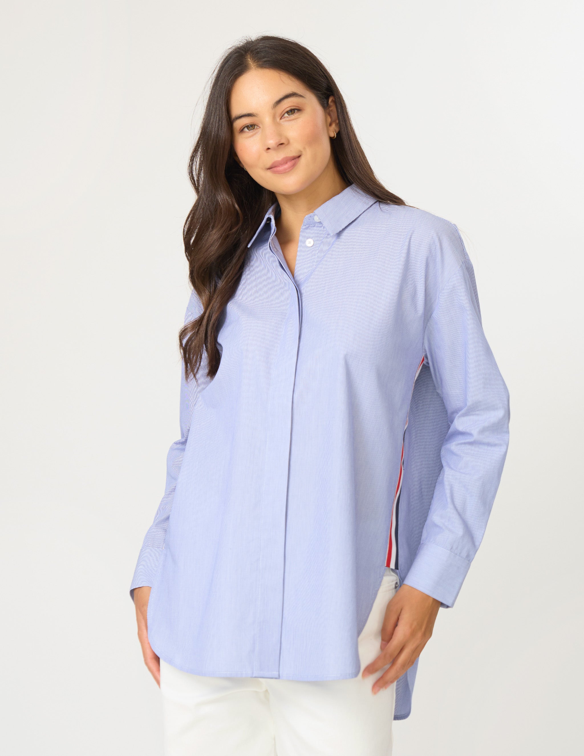 A woman modeling a light blue long-sleeve button-up shirt with subtle vertical textured stripes and red, white, and blue side panels, paired with white pants against a plain light gray background.