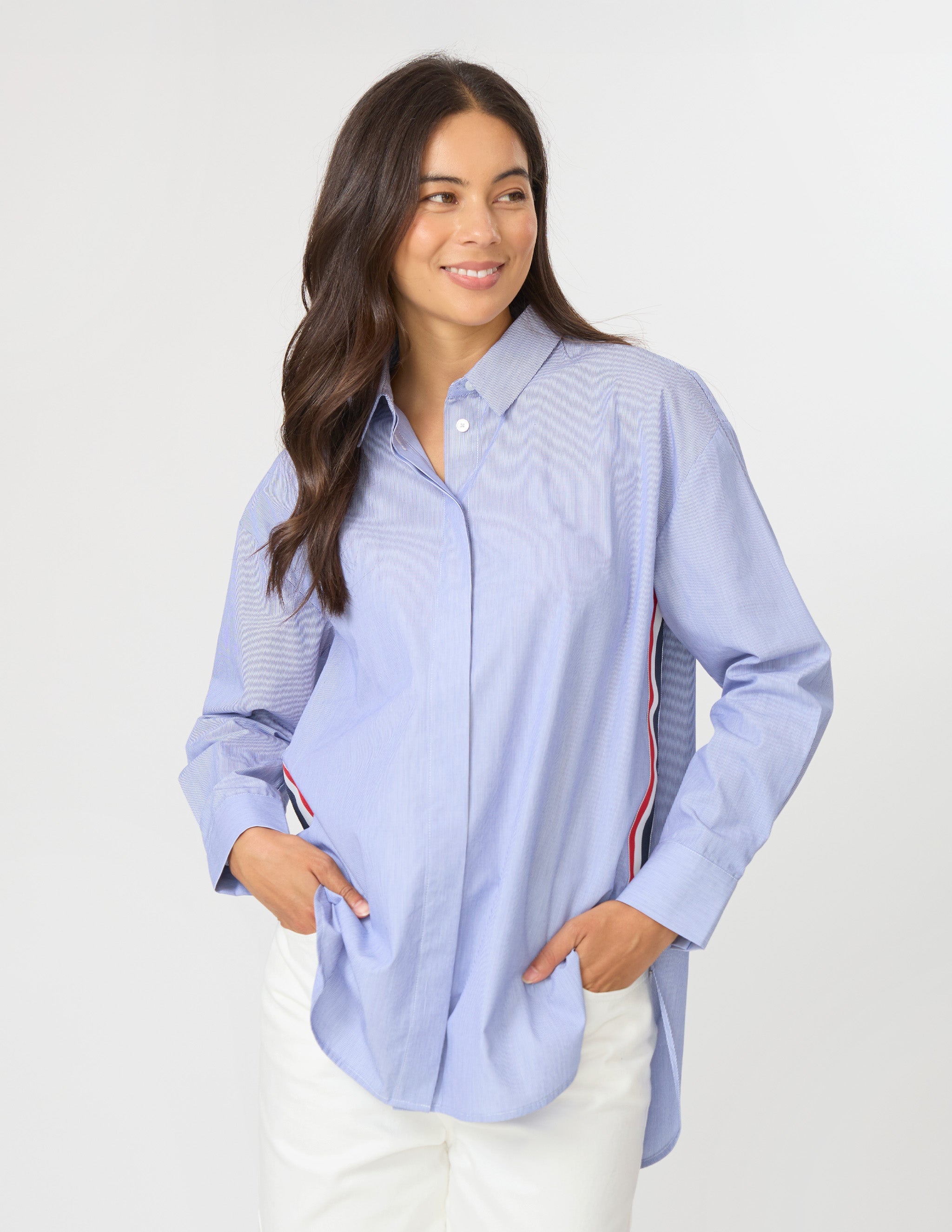 A woman modeling a light blue long-sleeve button-up shirt with subtle tonal wavy stripes and red and navy side stripes, paired with white pants against a light gray studio background.