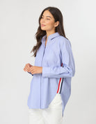 A woman wearing a light blue long-sleeve button-up shirt with a red, white, and navy stripe detail on the side, paired with white pants, against a plain light gray background.