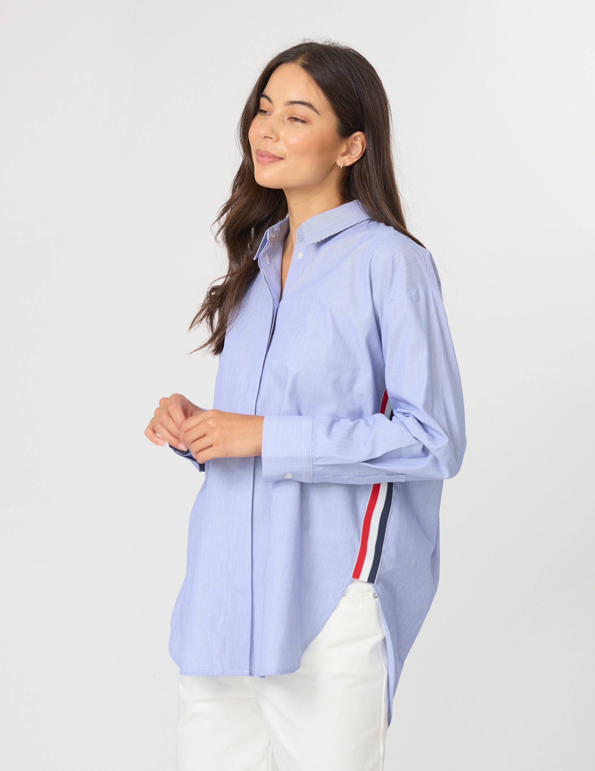 A woman wearing a light blue long-sleeve button-up shirt with a red, white, and navy stripe detail on the side, paired with white pants, against a plain light gray background.