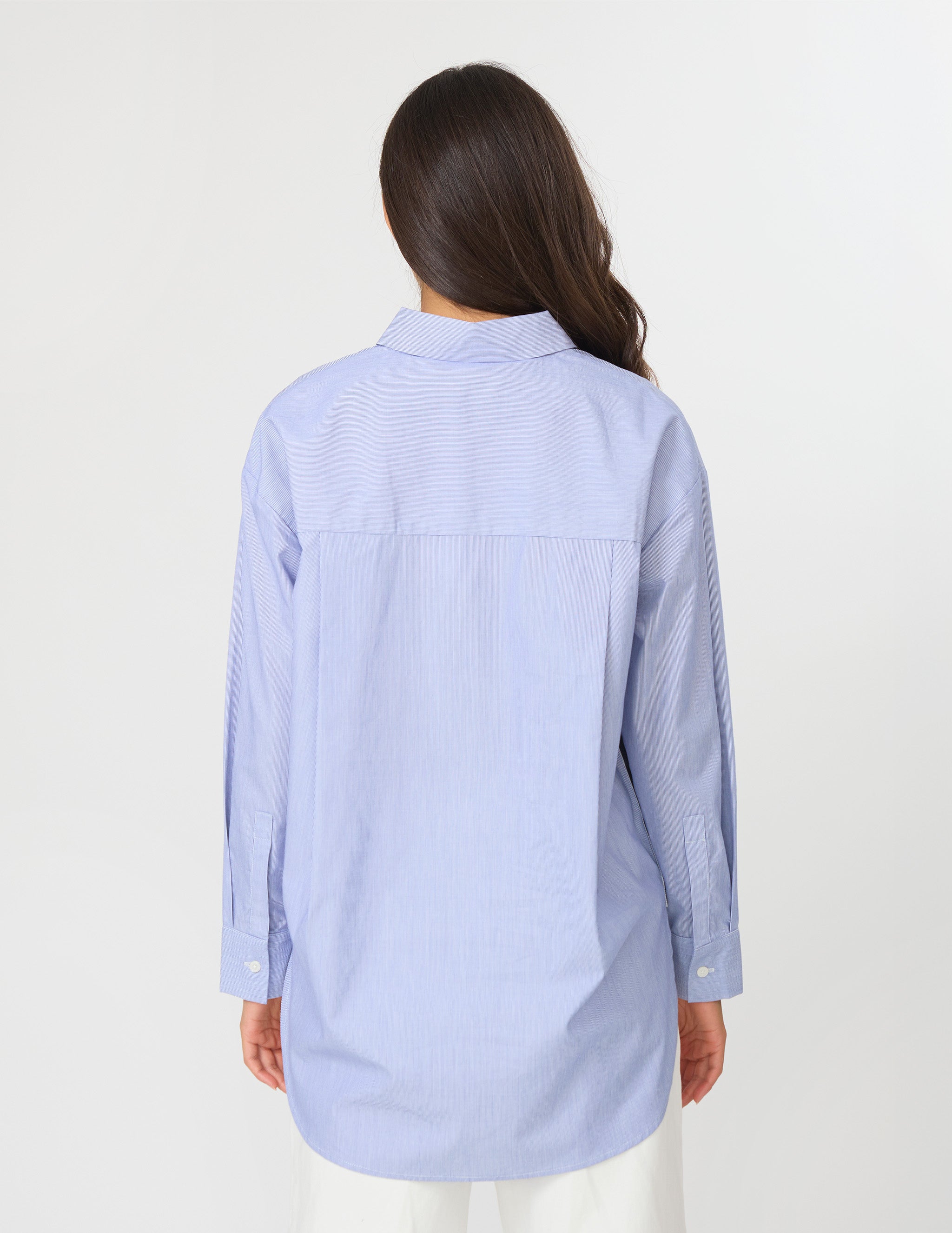 A woman viewed from the back wearing a light blue long-sleeve button-up shirt and white pants against a plain white studio background.
