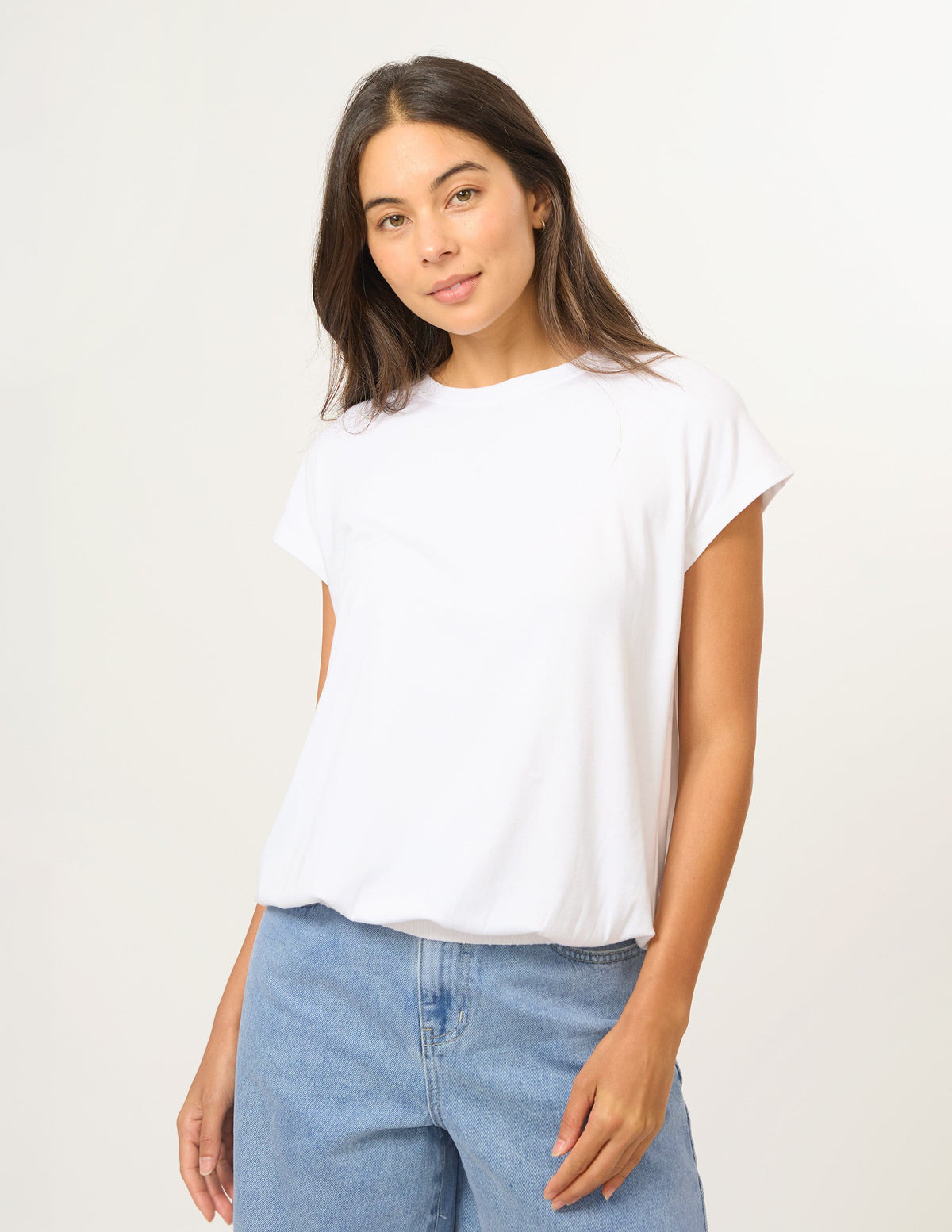 A woman wearing a white short-sleeve crewneck t-shirt and light blue jeans, standing against a plain off-white studio background.