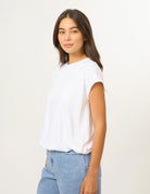 A woman wearing a white short-sleeve crewneck t-shirt and light blue jeans, standing against a plain off-white studio background.