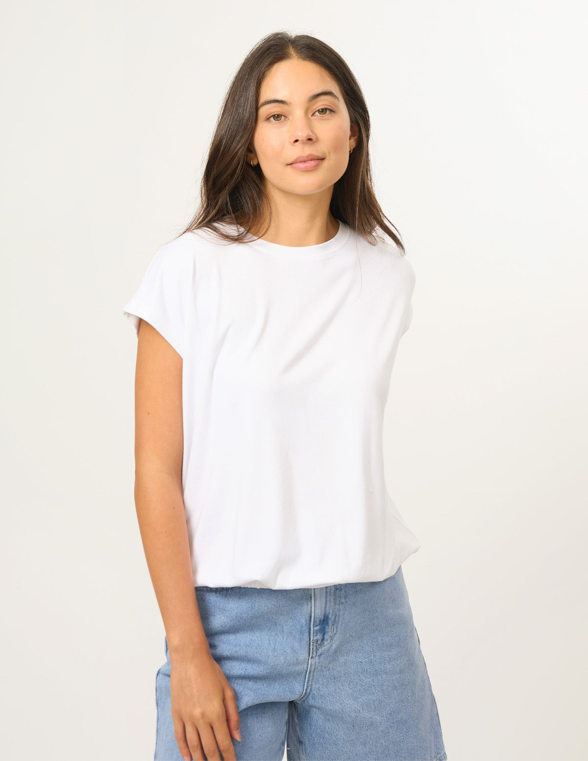 A woman modeling a white short-sleeve crewneck t-shirt paired with light blue jeans, standing against a plain off-white studio background.