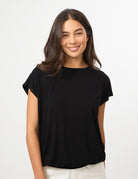 A woman with long dark brown hair wearing a black short-sleeve boat-neck top and white pants, standing against a plain white studio background.
