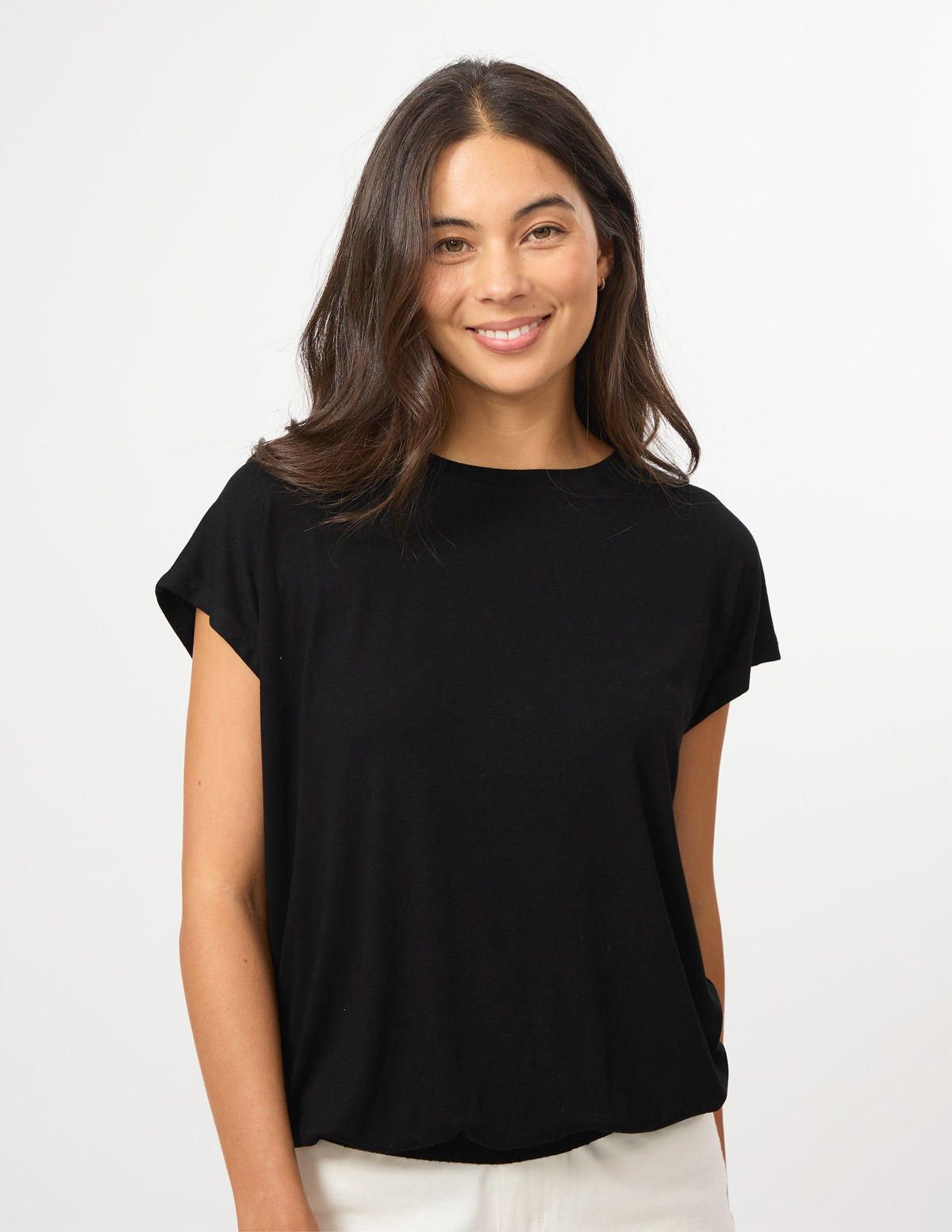 A woman with long dark brown hair wearing a black short-sleeve boat-neck top and white pants, standing against a plain white studio background.
