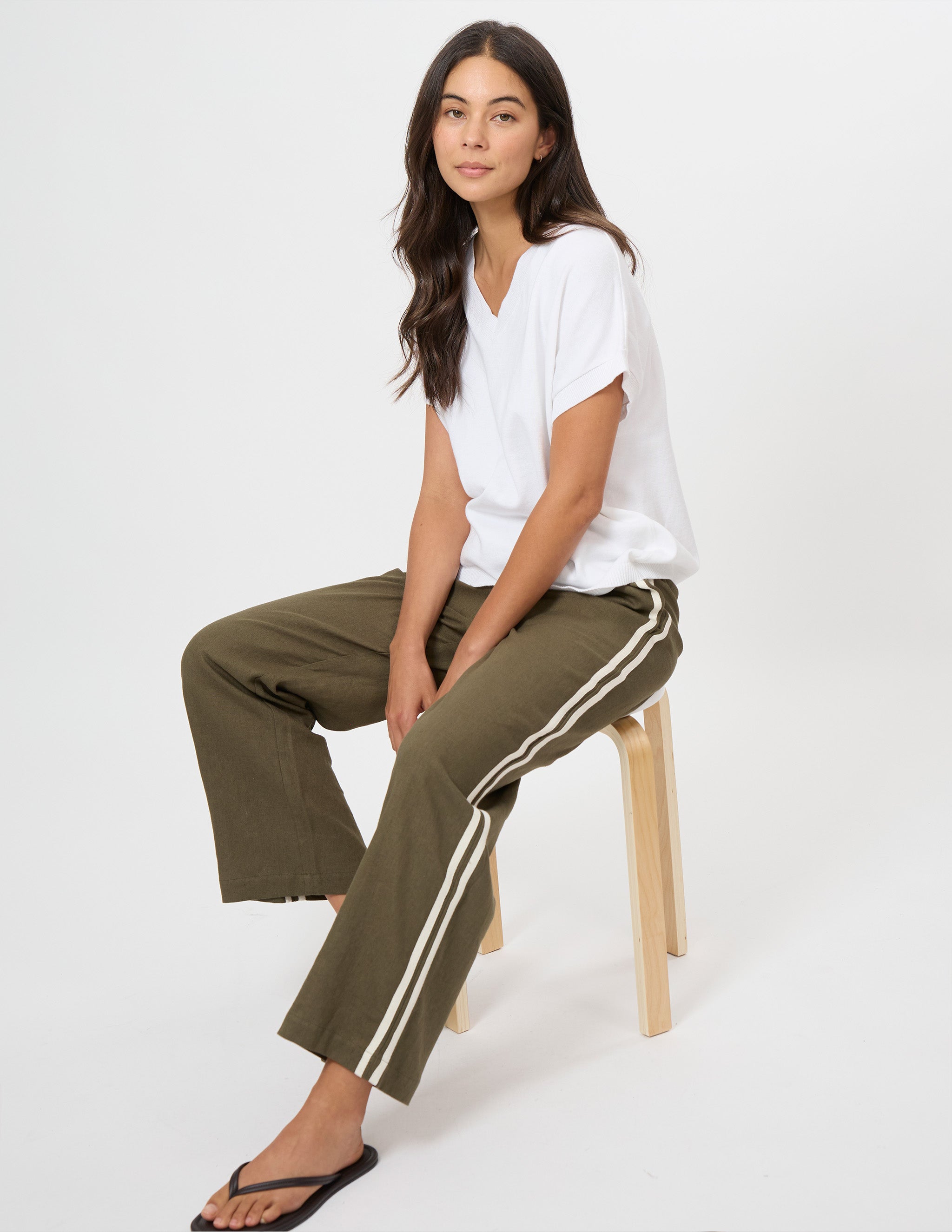 A woman sits on a light wood stool wearing olive green wide-leg pants with white side stripes, a white V-neck t-shirt, and black flip-flops against a plain white studio background.