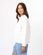 A woman wearing a white long-sleeve crewneck sweatshirt and light blue denim shorts, shown from a side-back angle against a plain white studio background.