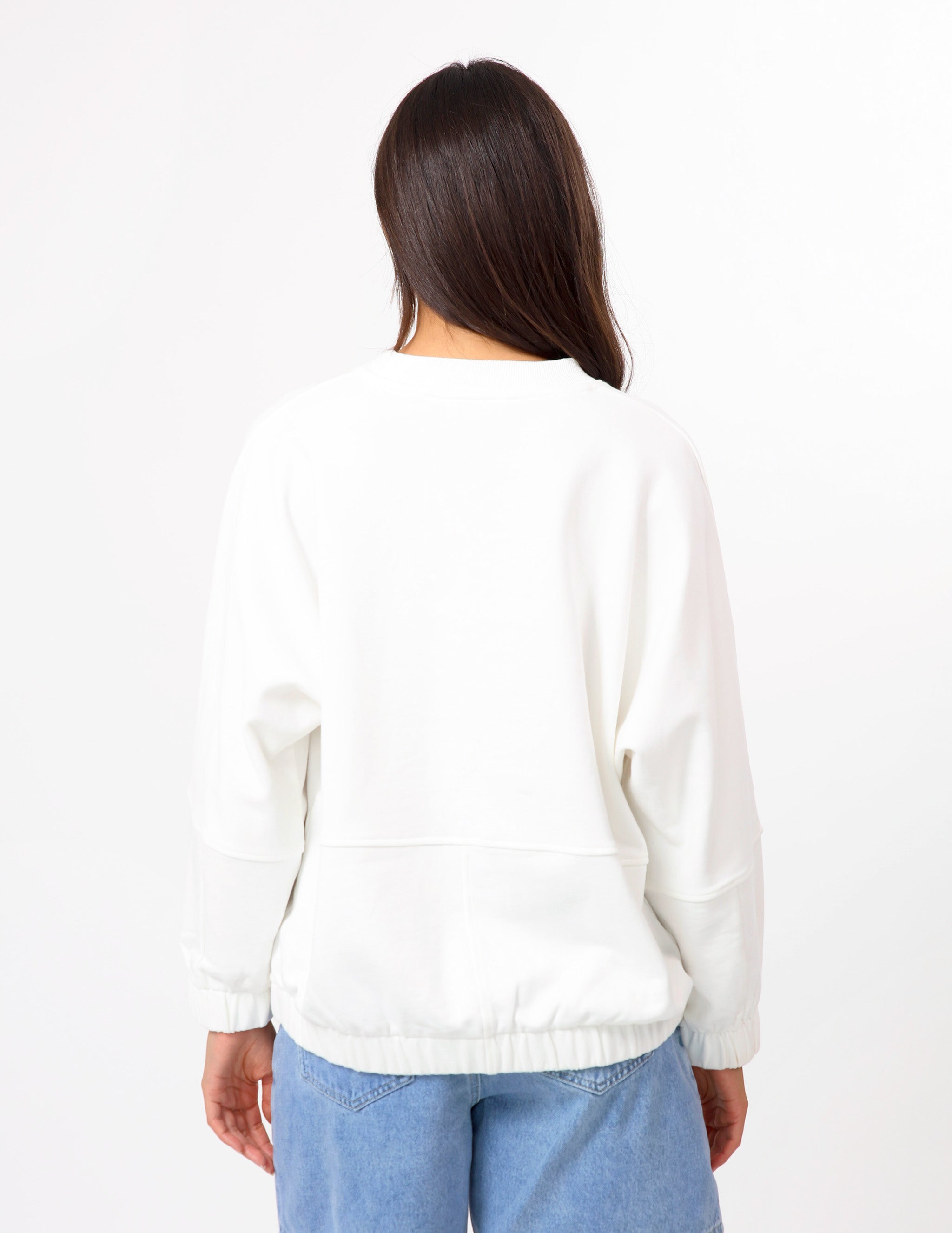 A woman viewed from behind wearing a white long-sleeve crewneck sweatshirt and light blue jeans against a plain white background.