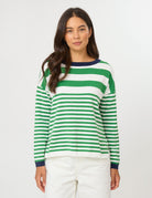 A woman wearing a green and white horizontally striped long-sleeve knit sweater with a navy blue crew neck, cuffs, and collar trim, paired with white pants, against a plain light gray studio background.