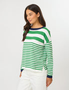 A woman wearing a green and white horizontally striped long-sleeve knit top with dark navy ribbed trim at the crew neck, cuffs, and hem, paired with white pants, against a light gray studio background.