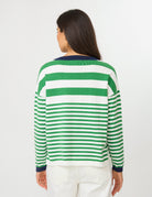 A woman viewed from the back wearing a green and white horizontally striped knit sweater with dark navy cuffs and collar, paired with white pants, against a plain light gray studio background.