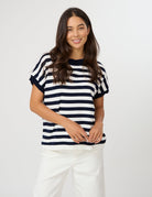 A woman wearing a navy and white horizontally striped short-sleeve knit top with solid navy trim, paired with white pants, photographed against a light gray studio background.