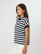 A woman wearing a navy and white horizontally striped short-sleeve knit top, paired with white pants, photographed against a plain light gray studio background.
