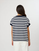 A woman viewed from the back wearing a short-sleeve striped T-shirt with navy and white horizontal stripes, paired with white pants, against a plain light gray studio background.