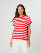 A woman modeling a red and white horizontally striped short-sleeve knit top with white pants, standing against a light gray studio background.