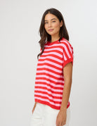 A woman wearing a red and white horizontally striped short-sleeve knit top with white pants, standing against a light gray studio background.