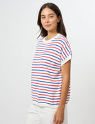 A woman modeling a short-sleeve knit top with horizontal red, blue, and white stripes, paired with white pants, against a plain light gray studio background.