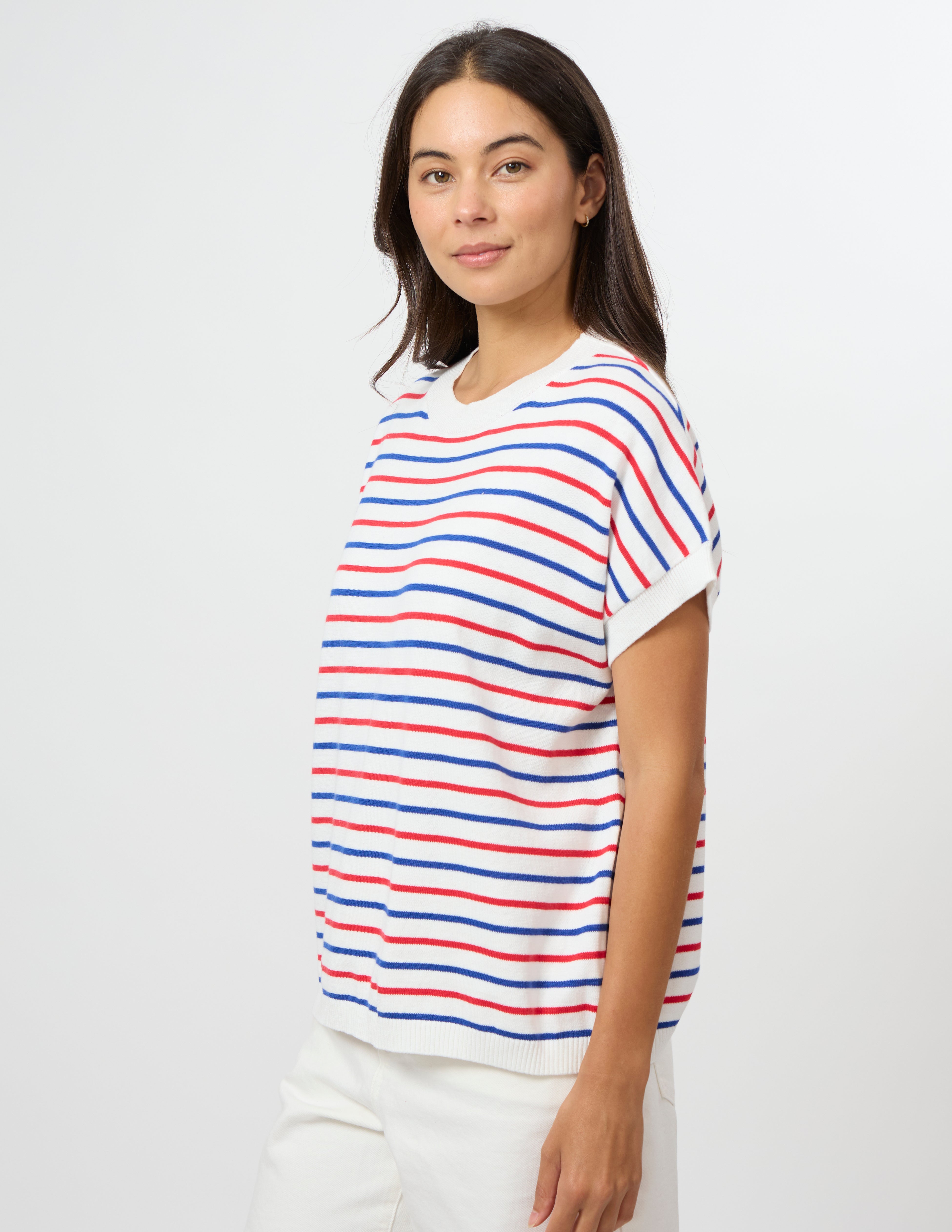 A woman modeling a short-sleeve knit top with horizontal red, blue, and white stripes, paired with white pants, against a plain light gray studio background.