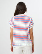 A woman viewed from the back wearing a white short-sleeve T-shirt with thin horizontal red and blue stripes, paired with white pants, against a plain light gray studio background.