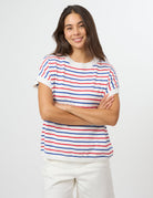A woman with long dark hair stands against a light gray studio background wearing a short-sleeve white knit top with thin red and blue horizontal stripes, paired with white pants.