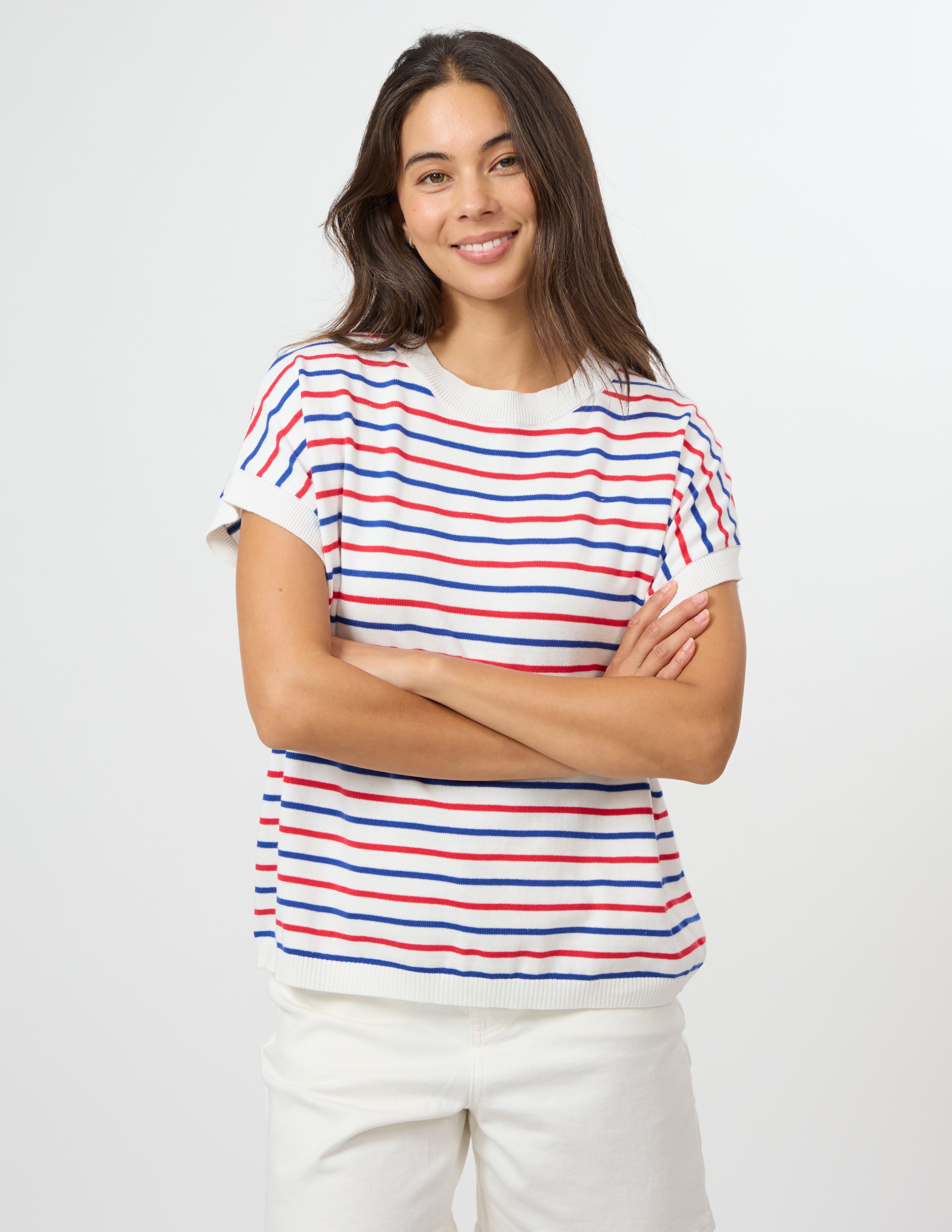 A woman with long dark hair stands against a light gray studio background wearing a short-sleeve white knit top with thin red and blue horizontal stripes, paired with white pants.