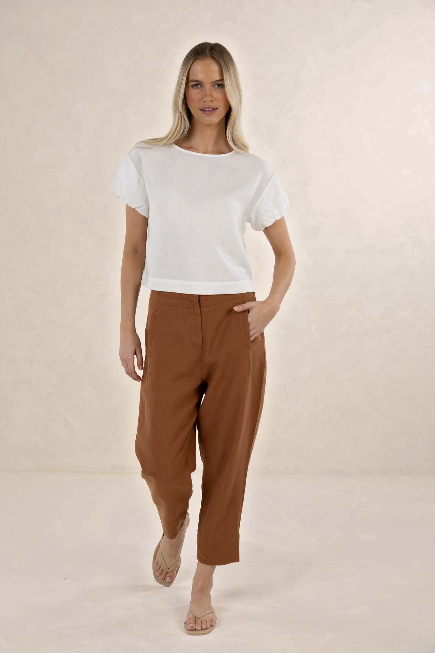 A woman modeling a white short-sleeve crop top and brown cropped trousers against a warm beige studio backdrop.