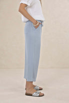 A model wearing light blue wide-leg cropped pants with a drawstring waist, paired with a white top and silver slide sandals, standing against a neutral beige background.
