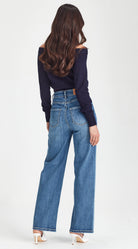 A woman shown from the back wearing high-waisted, wide-leg blue denim jeans and a dark navy off-the-shoulder long-sleeve top, paired with white high-heeled sandals, against a plain white studio background.