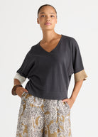A woman modeling a dark charcoal gray V-neck knit top with contrasting beige sleeve cuffs, paired with patterned trousers, against a plain light gray studio background.