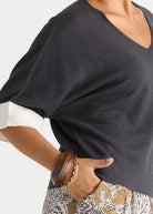Close-up of a person wearing a dark charcoal gray V-neck top with wide short sleeves, one rolled up to show a white undersleeve, paired with patterned pants and gold bracelets.