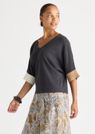A woman wearing a dark charcoal gray V-neck knit top with contrasting light beige cuffs and a patterned beige, white, and gray skirt, photographed against a light gray studio background.