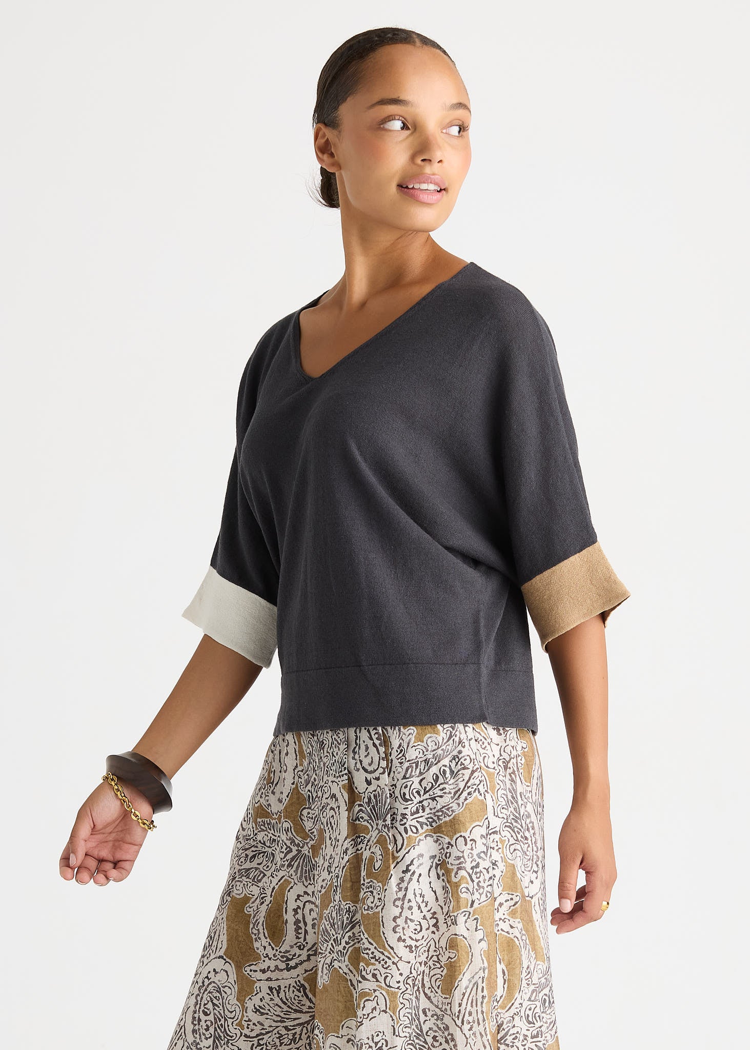 A woman wearing a dark charcoal gray V-neck knit top with contrasting light beige cuffs and a patterned beige, white, and gray skirt, photographed against a light gray studio background.